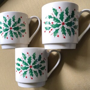May include: Three white ceramic mugs with a green holly and red berry design. The mugs are stacked on top of each other.