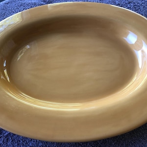 May include: A large, oval-shaped serving platter with a glossy, light brown finish. The ceramic dish has a wide, gently curved rim and a smooth, reflective surface. The platter is suitable for serving food.