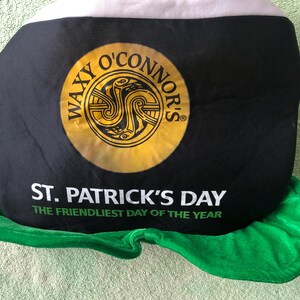 May include: A black and green felt hat with a white brim. The hat has a gold circle with the text "Waxy O'Connor's" and a Celtic knot design. The hat also has the text "St. Patrick's Day The Friendliest Day of the Year."