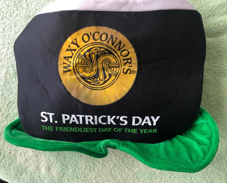 May include: A black and green felt hat with a white brim. The hat has a gold circle with the text "Waxy O'Connor's" and a Celtic knot design. The hat also has the text "St. Patrick's Day The Friendliest Day of the Year."