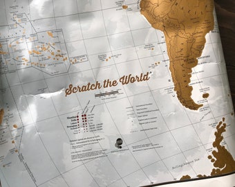 May include: A white scratch-off world map with gold foil accents. The map shows the continents of South America, Africa, and Australia. The text "Scratch the World" is printed in brown on the map. The map also includes a key with information about international boundaries, disputed boundaries, and highest peaks.