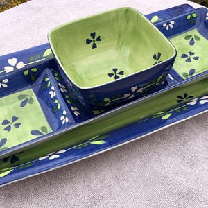 May include: A ceramic serving set with a rectangular tray and two square dishes, plus a square bowl. The set is painted in blue and green with white floral accents. The bowl and dishes have a green interior.