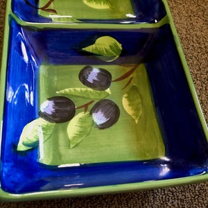 May include: A green and blue ceramic serving dish with a painted design of olive branches and olives. The dish has two compartments.