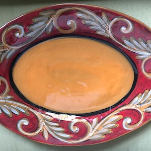 May include: An oval ceramic serving platter with a red and gold rim and a decorative design of gold and white leaves and swirls.