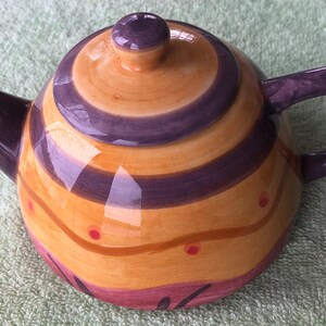 May include: A ceramic teapot with a vibrant design. The teapot features a yellow and purple spiral pattern, a purple handle, spout, and lid. Red dots and pink accents add to the decorative style. A charming addition to any kitchen.