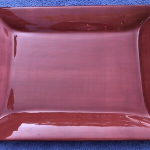 May include: A rectangular, burgundy-colored serving platter with a glossy finish. The edges are slightly curved, and the corners are rounded. The platter is empty and appears to be made of ceramic or a similar material. The background is a blue towel.