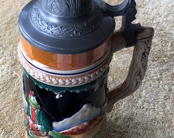 May include: A ceramic beer stein with a hinged pewter lid and handle. The stein features a detailed relief scene of a person in traditional attire in a mountain landscape. The base and rim are orange, with a brown and cream colored body.