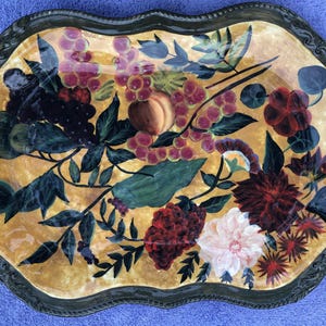 May include: An oval decorative serving tray with a gold-toned background and a dark, ornate border. The tray is adorned with a painted floral and fruit design, featuring grapes, peaches, and various flowers in shades of red, pink, and white.