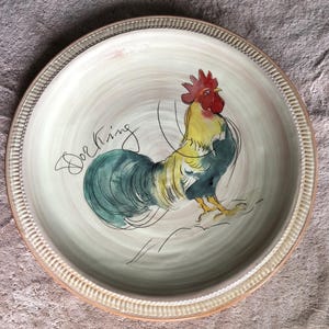 May include: A round ceramic plate with a cream-colored base and a textured, ridged rim. The center features a hand-painted illustration of a rooster with a red comb, yellow neck, and teal tail feathers, accompanied by the handwritten text Dorking.