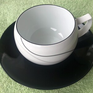 May include: A white teacup with a black rim and a thin black line design on the side. The teacup is sitting on a black saucer.