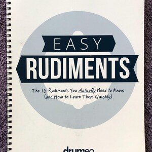 May include: A spiral-bound book titled "EASY RUDIMENTS" with text in a dark blue banner over a light blue circle. The text below reads "The 15 Rudiments You Actually Need to Know (and How to Learn Them Quickly)". The logo "drumeo" is at the bottom.