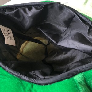 May include: Green plush hat with a black lining and a white label with the CE mark and text. The hat is turned inside out to show the lining.