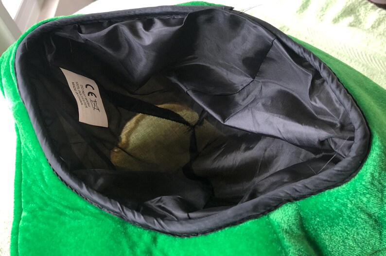 May include: Green plush hat with a black lining and a white label with the CE mark and text. The hat is turned inside out to show the lining.