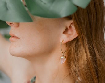 May include: Gold hoop earrings with a pink gemstone pendant. The earrings are hanging from a person's ear. The gemstone is a light pink color and has a gold cap.