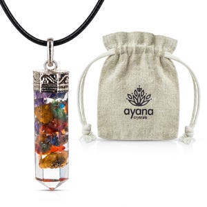 May include: A cylindrical pendant necklace filled with multicolored raw crystal chips, featuring a silver-toned metal cap and a black cord. Beside it sits a beige drawstring pouch with the printed text ayana crystals and a lotus icon.