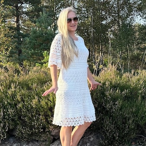 Op de afbeelding: Een witte gehaakte jurk met een kantversiering en korte mouwen. De jurk is knielengte en heeft een losse pasvorm. Het model draagt de jurk met witte hakken en een zonnebril.