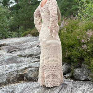 Könnte beinhalten: Ein langes, beiges Häkelkleid mit Rundhalsausschnitt und langen Ärmeln. Das Kleid hat eine lockere Passform und einen fließenden Rock.