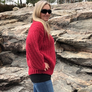May include: A woman wearing a red, chunky knit sweater, black top, and blue jeans. The sweater has a ribbed hem and cuffs. The woman is wearing sunglasses and is standing in front of a rocky background.