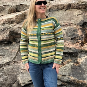 Op de afbeelding: Een groen, geel en wit gestreept gebreid vest met knoopsluiting. Het vest wordt gedragen door een persoon met lang blond haar en een zonnebril.