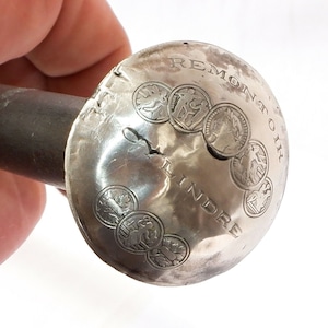 May include: A silver-toned metal knob with the words "Remontoir Lindre" engraved on it. The knob is decorated with a circle of five small, engraved images.