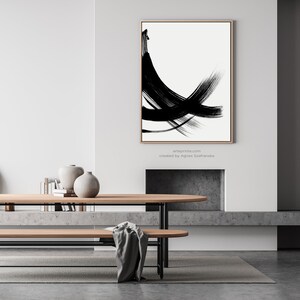 A black and white abstract painting with bold brushstrokes hangs above a wooden table and bench in a modern living room. The painting is framed.
Below the wall art, there is a text: "created by Agnes Szafranska - arteprinte.com".