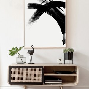 A black and white abstract minimalist art print in a wooden frame hangs above a wooden cabinet with a black and woven rattan door. A black bird figurine and a potted plant are on top of the cabinet.
Over the wall art, there is a text: "created by Agnes Szafranska - arteprinte.com".