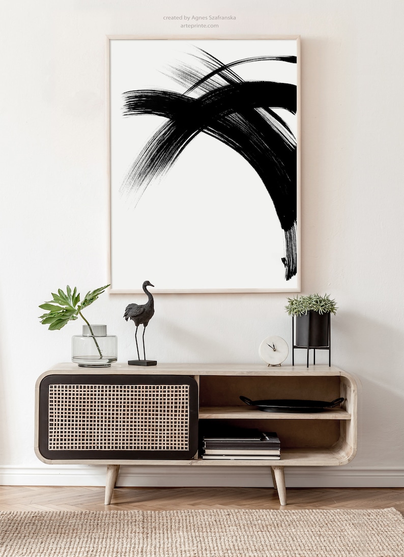 A black and white abstract minimalist art print in a wooden frame hangs above a wooden cabinet with a black and woven rattan door. A black bird figurine and a potted plant are on top of the cabinet.
Over the wall art, there is a text: "created by Agnes Szafranska - arteprinte.com".