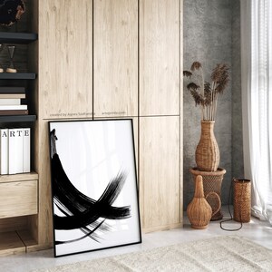 A black and white abstract art print with bold brushstrokes in a black frame standing on a floor in a room with wooden cabinets and a woven vase with dried flowers.
Over the wall art, there is a text: "created by Agnes Szafranska - arteprinte.com".