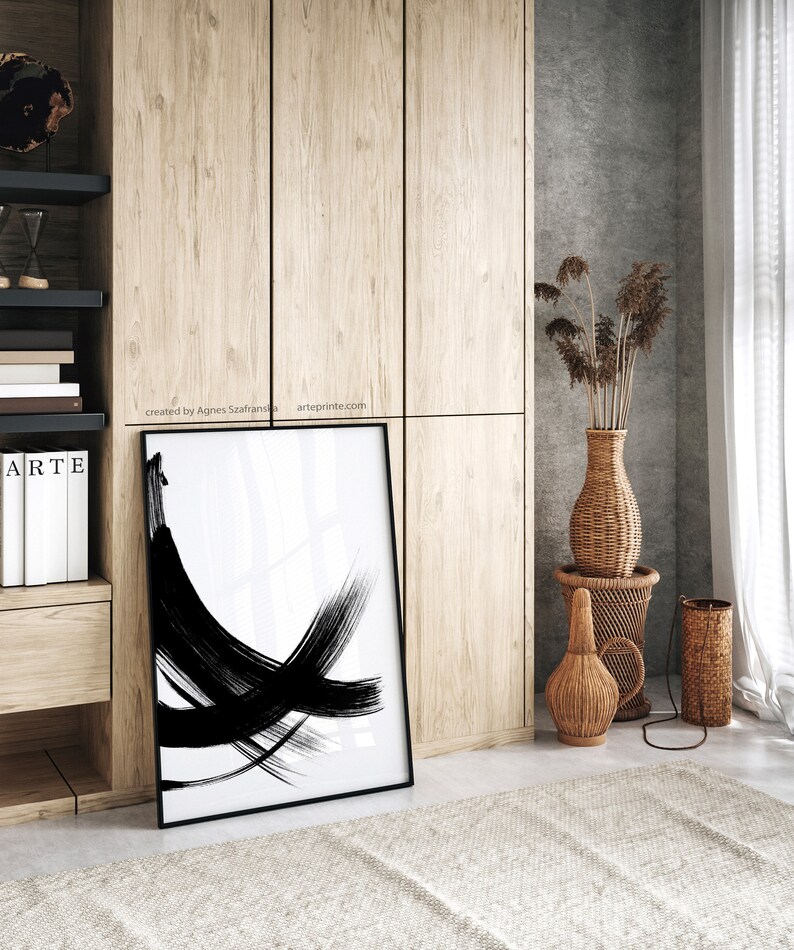 A black and white abstract art print with bold brushstrokes in a black frame standing on a floor in a room with wooden cabinets and a woven vase with dried flowers.
Over the wall art, there is a text: "created by Agnes Szafranska - arteprinte.com".