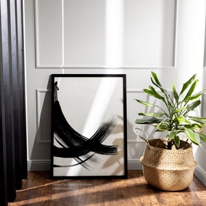 Black and white abstract art print in a black frame, standing on a wooden floor  with a potted plant in a woven basket in the corner.
Below the wall art, there is a text: "created by Agnes Szafranska - arteprinte.com".