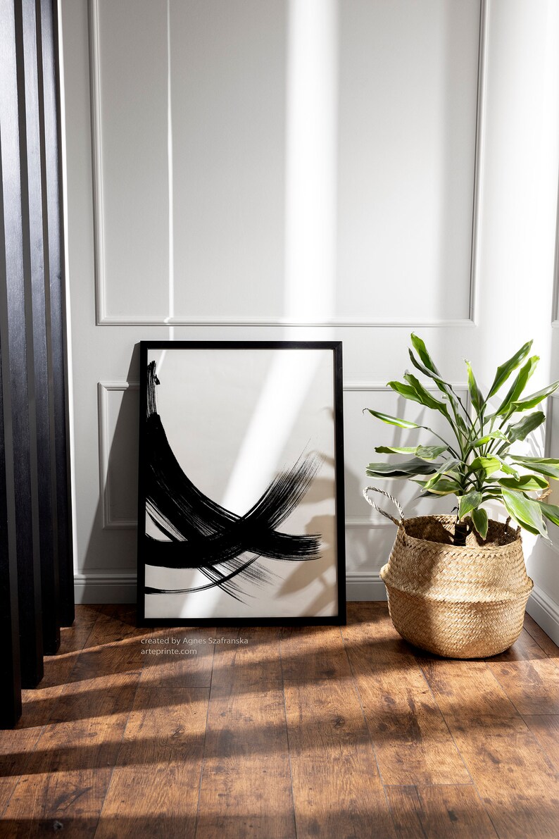 Black and white abstract art print in a black frame, standing on a wooden floor  with a potted plant in a woven basket in the corner.
Below the wall art, there is a text: "created by Agnes Szafranska - arteprinte.com".