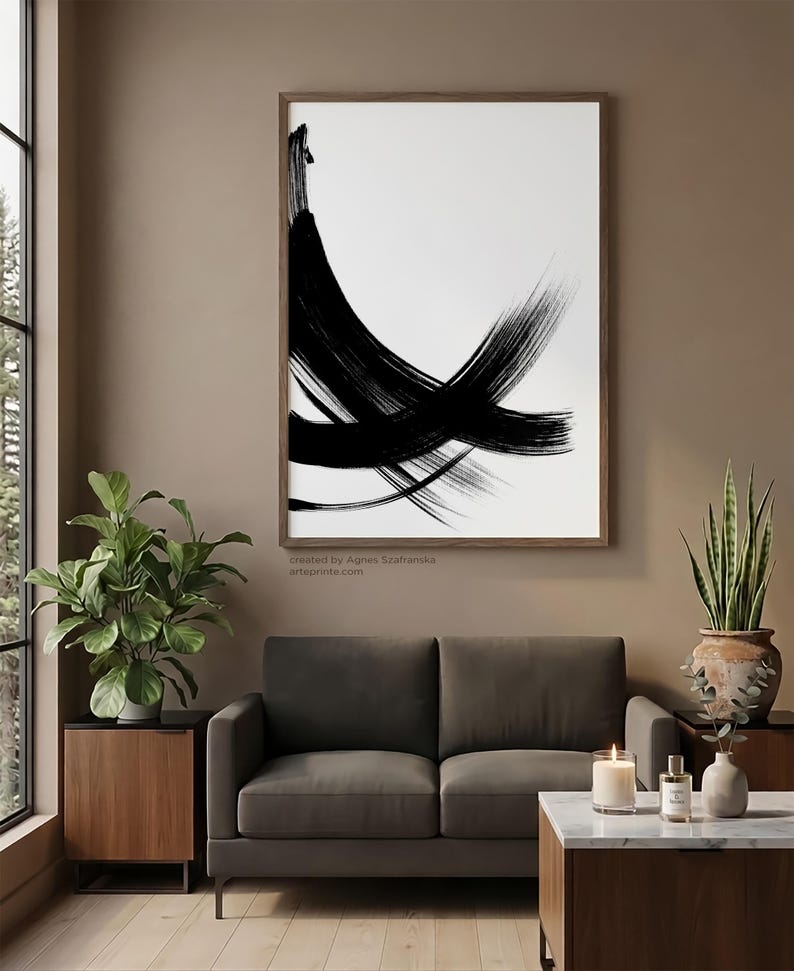 Vertical wall art in a warm mid-century modern living room featuring a large wood-framed black and white minimalist line art print above a brown sofa.
The interior includes a fiddle leaf fig, a snake plant in a rustic pot, and a marble coffee table with a candle. Large window with a pine forest view creates a bright, natural atmosphere.
Below the artwork, there is a text: "created by Agnes Szafranska - arteprinte.com".