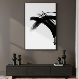 A large black and white minimalist abstract ink painting in a sleek black frame, mounted on a dark warm grey wall. The artwork is styled above a modern dark wood sideboard decorated with contemporary black vases and books, creating a sophisticated and moody Japandi interior.
Below the wall art, there is a text: "created by Agnes Szafranska - arteprinte.com".