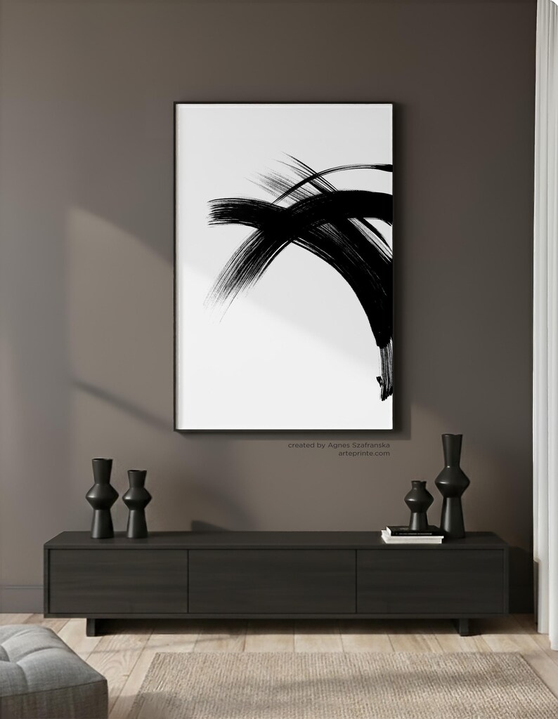 A large black and white minimalist abstract ink painting in a sleek black frame, mounted on a dark warm grey wall. The artwork is styled above a modern dark wood sideboard decorated with contemporary black vases and books, creating a sophisticated and moody Japandi interior.
Below the wall art, there is a text: "created by Agnes Szafranska - arteprinte.com".