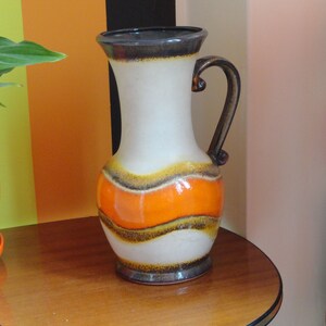 May include: A white ceramic vase with brown and orange stripes. The vase has a wide mouth and a handle. It is sitting on a wooden table.