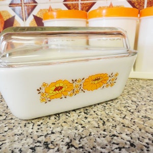 May include: White ceramic casserole dish with a glass lid and a floral design featuring orange and brown flowers. The dish is sitting on a worktop.