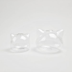 May include: Two clear glass vases shaped like cat heads. The larger vase is in the background and the smaller vase is in the foreground.