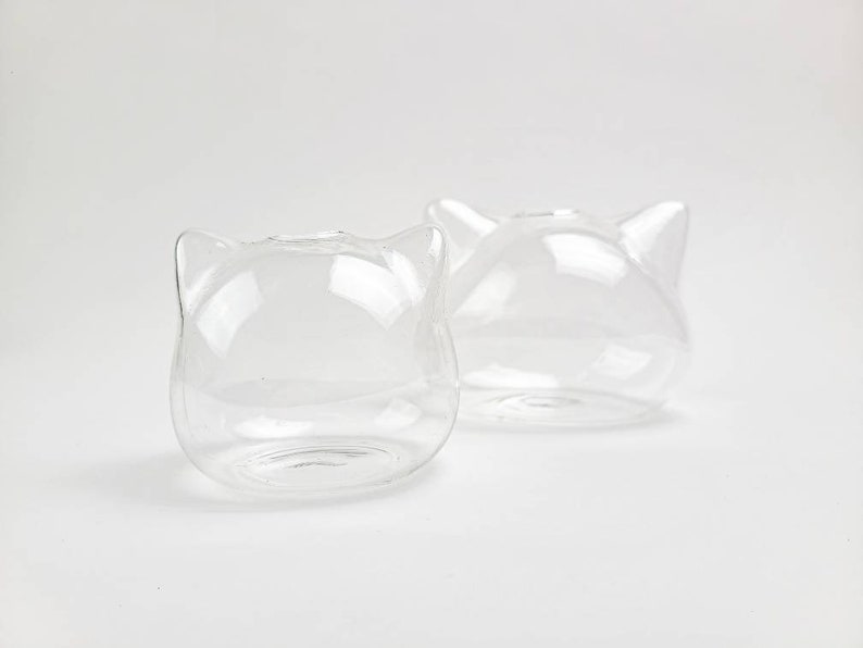 May include: Two clear glass cat-shaped vases. The vases are empty and have a smooth, rounded surface.