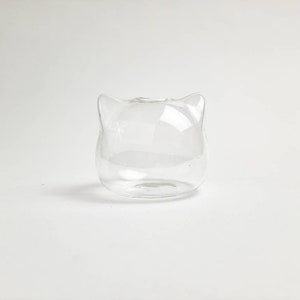 May include: A clear glass vase shaped like a cat's head. The vase has two small ears on top and a round body.
