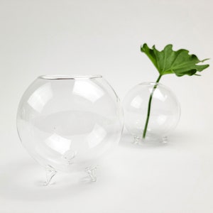 May include: Two clear, spherical glass vases with three small legs. One vase holds a green leaf. The vases are transparent, allowing light to pass through. The design is minimalist and modern, suitable for displaying small plants or flowers.