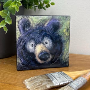May include: A square ceramic tile with a cartoon-style illustration of a black bear with large eyes and a light brown snout. The bear is set against a green and yellow background. Two paint brushes are in the foreground.