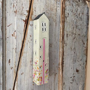 May include: A tall, slender, off-white ceramic house sculpture with a black peaked roof. The facade features hand-painted black outlines of small square windows, a large double window, and a vertical pink stripe. The base is decorated with colourful dots.