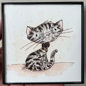 May include: A framed square print with a cartoon-style illustration of a grey tabby cat. The cat has a large head and wears a collar with a gold charm. The background features a soft, pink watercolour wash.