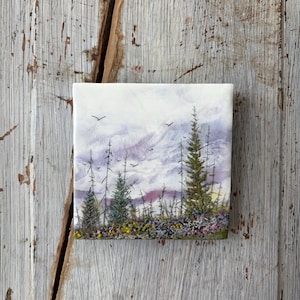 May include: A square ceramic tile featuring a painted landscape of evergreen trees and a field of wildflowers under a cloudy, light purple and white sky. Several small black birds are depicted flying in the distance above the forest.