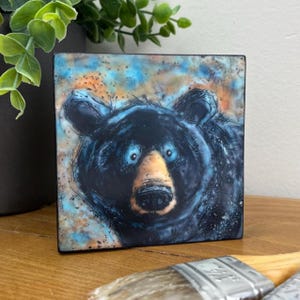 May include: Square ceramic tile featuring a black bear with blue eyes and a tan snout. The bear is set against a background of blue and orange hues. The artwork has a painterly, sketched style.