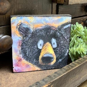 May include: Square wooden block with a cartoon-style illustration of a black bear's face. The bear has large blue eyes, a yellow nose, and a black outline. The background features an abstract design with orange, purple, and blue colours.