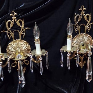 May include: Two decorative gold-coloured wall sconces with crystal pendants and candle-shaped light bulbs. Each sconce has an ornate backplate and detailed design, creating a vintage aesthetic.