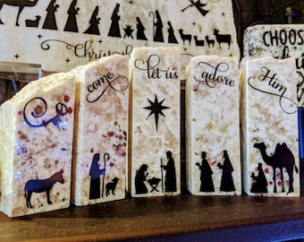 Puede incluir: Conjunto de seis pequeños bloques de piedra rectangulares de color beige con letras e imágenes negras. Los bloques presentan mensajes e imágenes con temática navideña, incluyendo "Let us adore Him", "Come", "Let us adore" y una estrella.