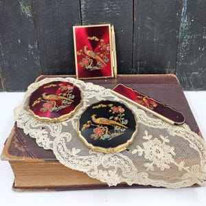 May include: A vintage vanity set featuring a red compact, two round compacts, and a comb, all with gold trim. Each piece is adorned with a detailed pheasant and floral design. The set is displayed on a lace doily.