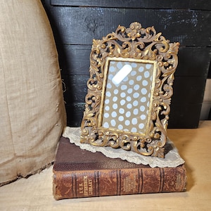 May include: An ornate, rectangular picture frame with a distressed gold finish and intricate scrollwork detailing. The frame features a central glass pane displaying a tan background with a repeating pattern of white, irregular circular dots.